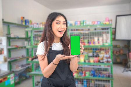 Asian female work as groceries store or modern store staff pointing to the copy space at smartphone blank screen, wearing black apron while standing in front of display rackの写真素材
