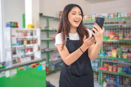Asian female work as groceries store or modern store staff pointing to the copy space at smartphone blank screen, wearing black apron while standing in front of display rackの写真素材