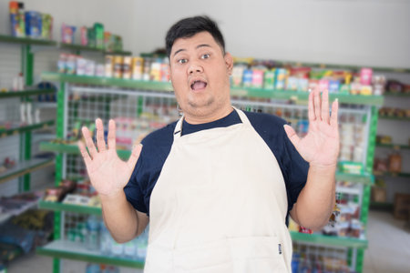Young Asian man as grocery store staff standing surprise pose with grey apron in front of display shelvesの写真素材