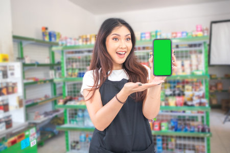 Asian female work as groceries store or modern store staff pointing to the copy space at smartphone blank screen, wearing black apron while standing in front of display rackの写真素材