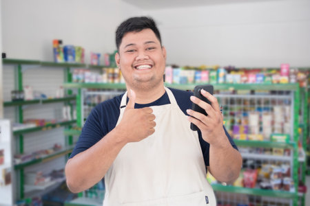 Asian male work as groceries store staff smille giving gesturing thumbs up for approval and looks at the phone, wearing grey apron while standing in front of display rackの写真素材