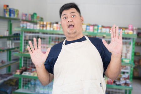 Young Asian man as grocery store staff standing surprise pose with grey apron in front of display shelvesの写真素材