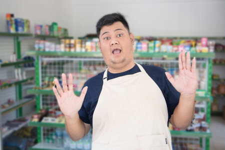 Young Asian man as grocery store staff standing surprise pose with grey apron in front of display shelvesの写真素材