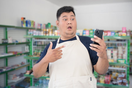 Asian male work as groceries store staff surprised, shocked and looks at the phone, wearing grey apron while standing in front of display rackの写真素材