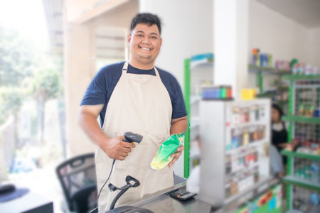 Smiling young Asian man as grocery store staff standing at cashier counter, cashier scanning grocery items and full of confidence in front of display shelvesの写真素材