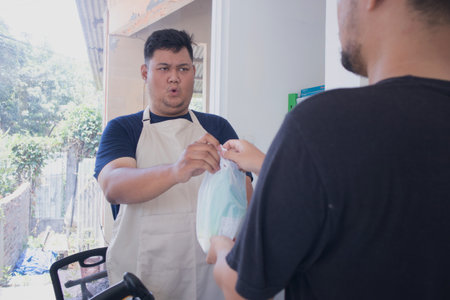 young asian man cashier worker standing near cashier counter in grey and making payment and giving goods to customer in grocery store or modern marketの写真素材