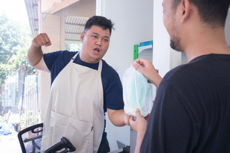 young asian male cashier worker standing near gray cashier counter about to hit customer for not paying in grocery store or modern marketの写真素材