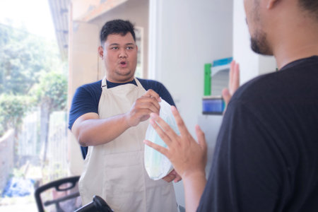 young asian man cashier worker standing near cashier counter in grey and making payment and giving goods to customer in grocery store or modern marketの写真素材