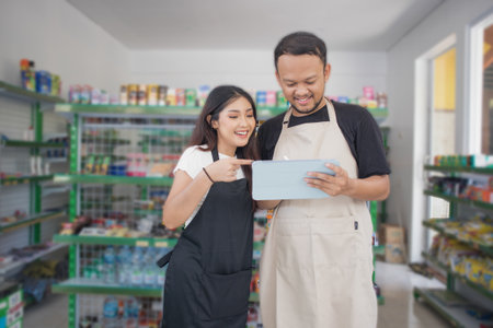 friendly Asian Cashier couple discussing pointing tab, cashier wearing black and beige apron standing in grocery or convenience storeの写真素材