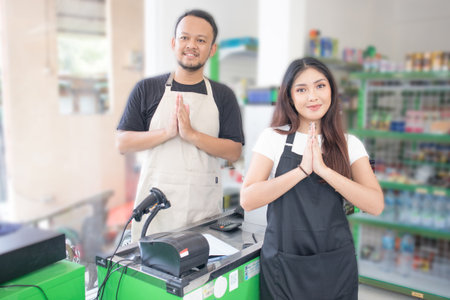 couple Friendly Asian cashier welcoming customer with open arms, cashier is wearing black and cream apron standing in a groceries or convenient storeの写真素材