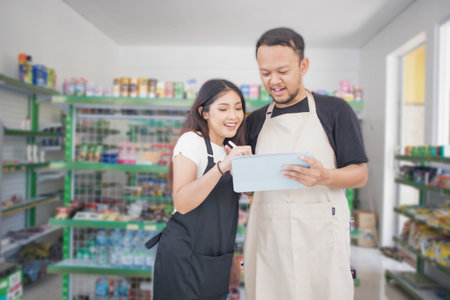 friendly Asian Cashier couple discussing pointing tab, cashier wearing black and beige apron standing in grocery or convenience storeの写真素材