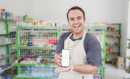 Asian male work as groceries store or modern store staff pointing to the copy space at smartphone blank screen, wearing grey apron while standing in front of display rackの写真素材