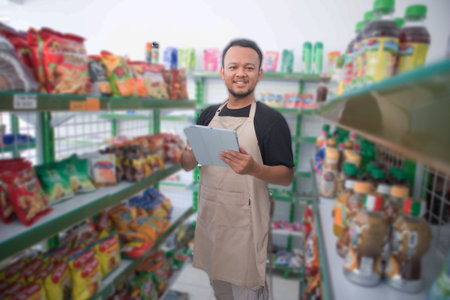 Happy Asian man as groceries or modren market staff checking products and goods on the display shelf while holding tablet and wearing grey apronの写真素材