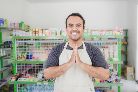 Young smiling Asian man as grocery store staff standing at cashier counter with arms namaste pose and full of confidence in front of display shelvesの写真素材