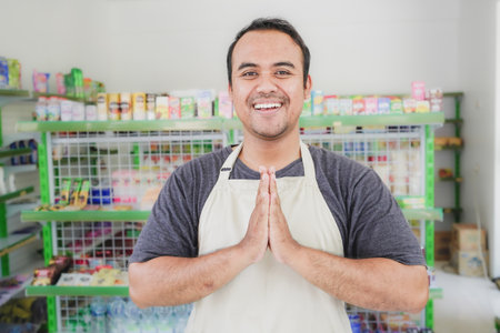 Young smiling Asian man as grocery store staff standing at cashier counter with arms namaste pose and full of confidence in front of display shelvesの写真素材
