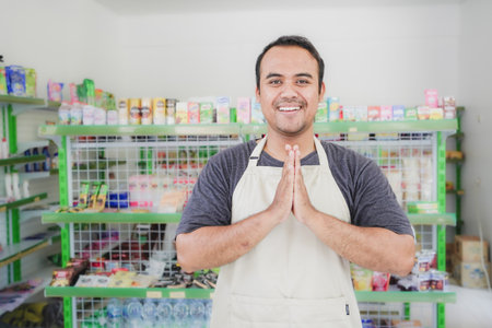 Young smiling Asian man as grocery store staff standing at cashier counter with arms namaste pose and full of confidence in front of display shelvesの写真素材
