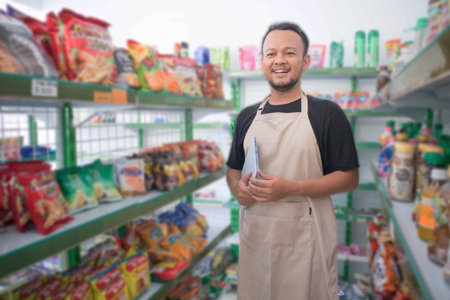 Happy Asian man as groceries or modren market staff checking products and goods on the display shelf while holding tablet and wearing grey apronの写真素材