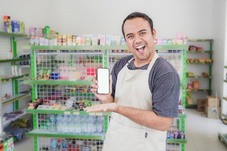 Asian male work as groceries store or modern store staff pointing to the copy space at smartphone blank screen, wearing grey apron while standing in front of display rackの写真素材