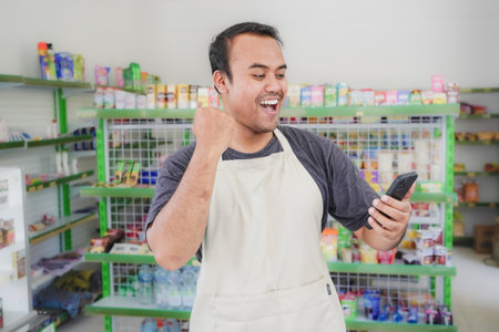 Asian male work as groceries store or modern market staff smiles , happy looks at the phone, wearing...