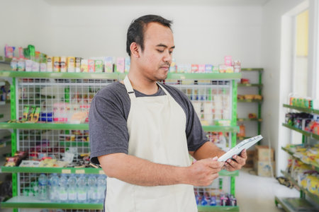 Happy Asian man as groceries or modern market staff checking products and goods on the display shelf while holding tablet and wearing grey apron in front of display shelvesの写真素材