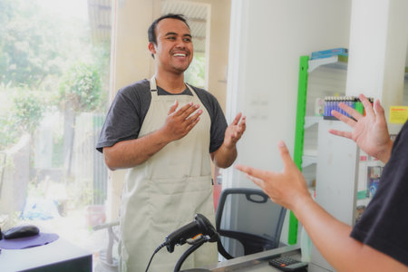young asian man cashier worker standing near cashier counter in black apron and making payment and giving goods to customer in grocery store or modern marketの写真素材