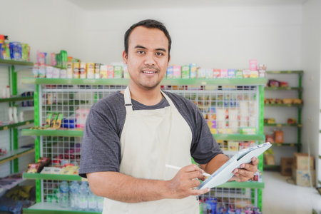 Happy Asian man as groceries or modern market staff checking products and goods on the display shelf while holding tablet and wearing grey apron in front of display shelvesの写真素材