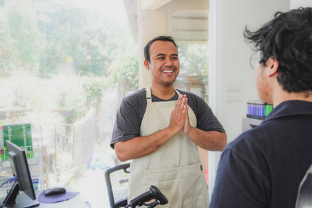 young asian man cashier worker standing near cashier counter in black apron and making payment and giving goods to customer in grocery store or modern marketの写真素材