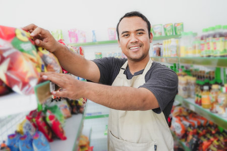 Smiling young Asian man at a modern grocery store or market wearing a gray apron checking goods on a display shelfの写真素材