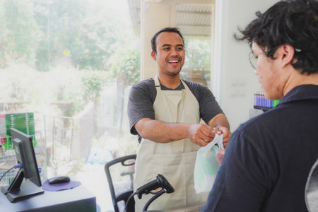 young asian man cashier worker standing near cashier counter in black apron and making payment and giving goods to customer in grocery store or modern marketの写真素材