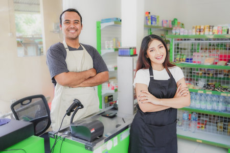 couple Friendly Asian cashier welcoming customer with open arms, cashier is wearing black and cream apron standing in a groceries or convenient storeの写真素材