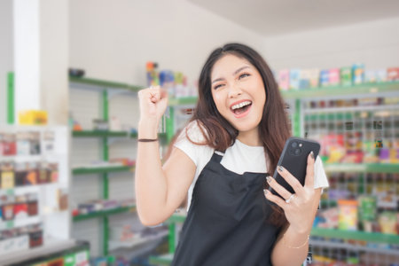 Asian female work as groceries store staff smiles and looks at the phone, wearing black apron while...