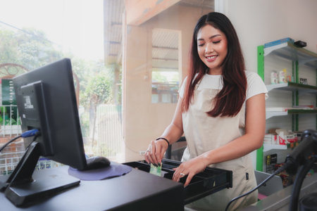 Smiling young Asian woman, grocery store staff wearing apron standing at cashier counter, returning change to buyerの写真素材