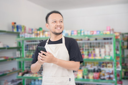 Asian male work as groceries store or modern market staff smiles and looks at the phone, wearing cream apron while standing in front of display rackの写真素材