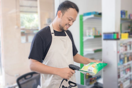 Smiling young Asian man as grocery store staff standing at cashier counter, cashier scanning grocery items and full of confidence in front of display shelvesの写真素材