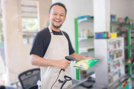 Smiling young Asian man as grocery store staff standing at cashier counter, cashier scanning grocery items and full of confidence in front of display shelvesの写真素材