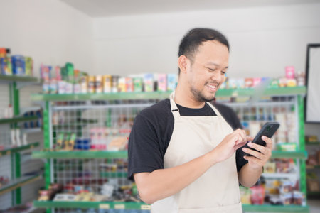 Asian male work as groceries store or modern market staff smiles and looks at the phone, wearing cream apron while standing in front of display rackの写真素材
