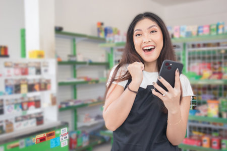 Asian female work as groceries store staff smiles and looks at the phone, wearing black apron while standing in front of display rackの写真素材