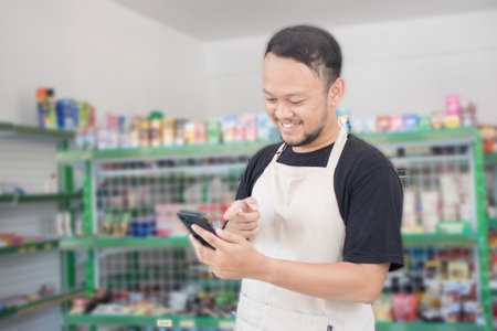 Asian male work as groceries store or modern market staff smiles and looks at the phone, wearing cream apron while standing in front of display rackの写真素材