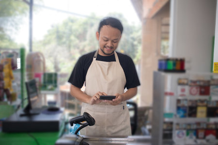 Smiling young Asian man as grocery store staff standing at cashier counter, cashier scanning grocery items and full of confidence in front of display shelvesの写真素材