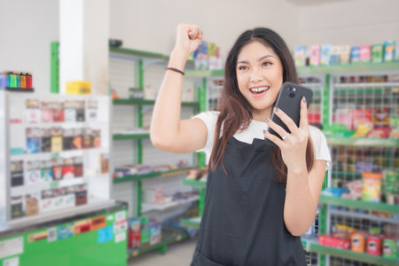 Asian female work as groceries store staff smiles and looks at the phone, wearing black apron while standing in front of display rackの写真素材