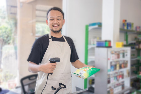 Smiling young Asian man as grocery store staff standing at cashier counter, cashier scanning grocery...