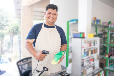 Smiling young Asian man as grocery store staff standing at cashier counter, cashier scanning grocery items and full of confidence in front of display shelvesの写真素材