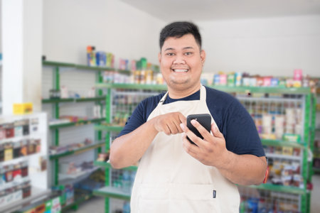 Asian male works as grocery store staff, smiling while looking at the phone, wearing a white apron while standing in front of display racks.の写真素材