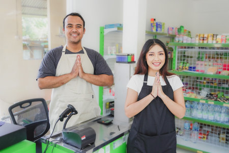 couple Asian cashier welcoming customer with open arms, cashier is wearing black and cream apron standing in a groceries or convenient storeの写真素材