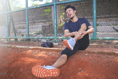 A male runner, sportsman sits on the roadside in sportswear and holds his leg, feels pain in his...