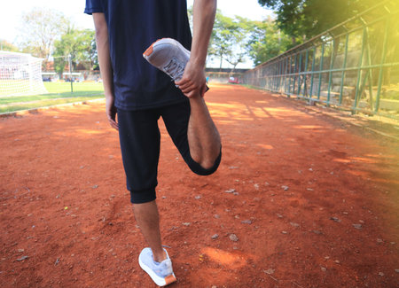 A man is stretching his leg on a dirt field. The man is wearing a grey shirt and black shorts