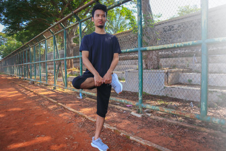A man is stretching his leg on a dirt field. The man is wearing a black shirt and black shorts