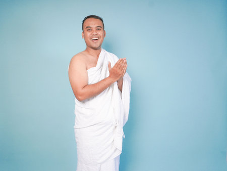 young Asian Muslim man wearing ihram posing praying and lifting head up over isolated blue...