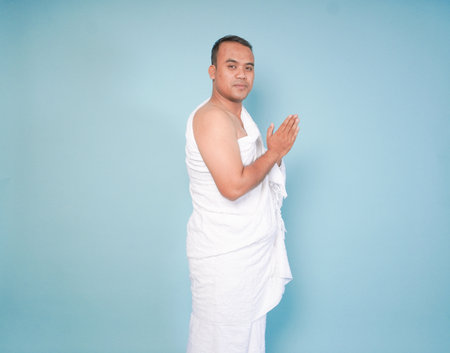 young Asian Muslim man wearing ihram gesturing, pose traditional religious greeting, isolated by...