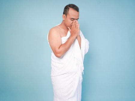 young Asian Muslim man wearing ihram posing praying and lifting head up over isolated blue...